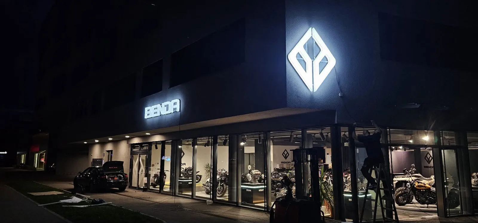Benda Motor bei Nacht: beleuchtetes Logo und Schriftzug an der Fassade über der Glasfront.