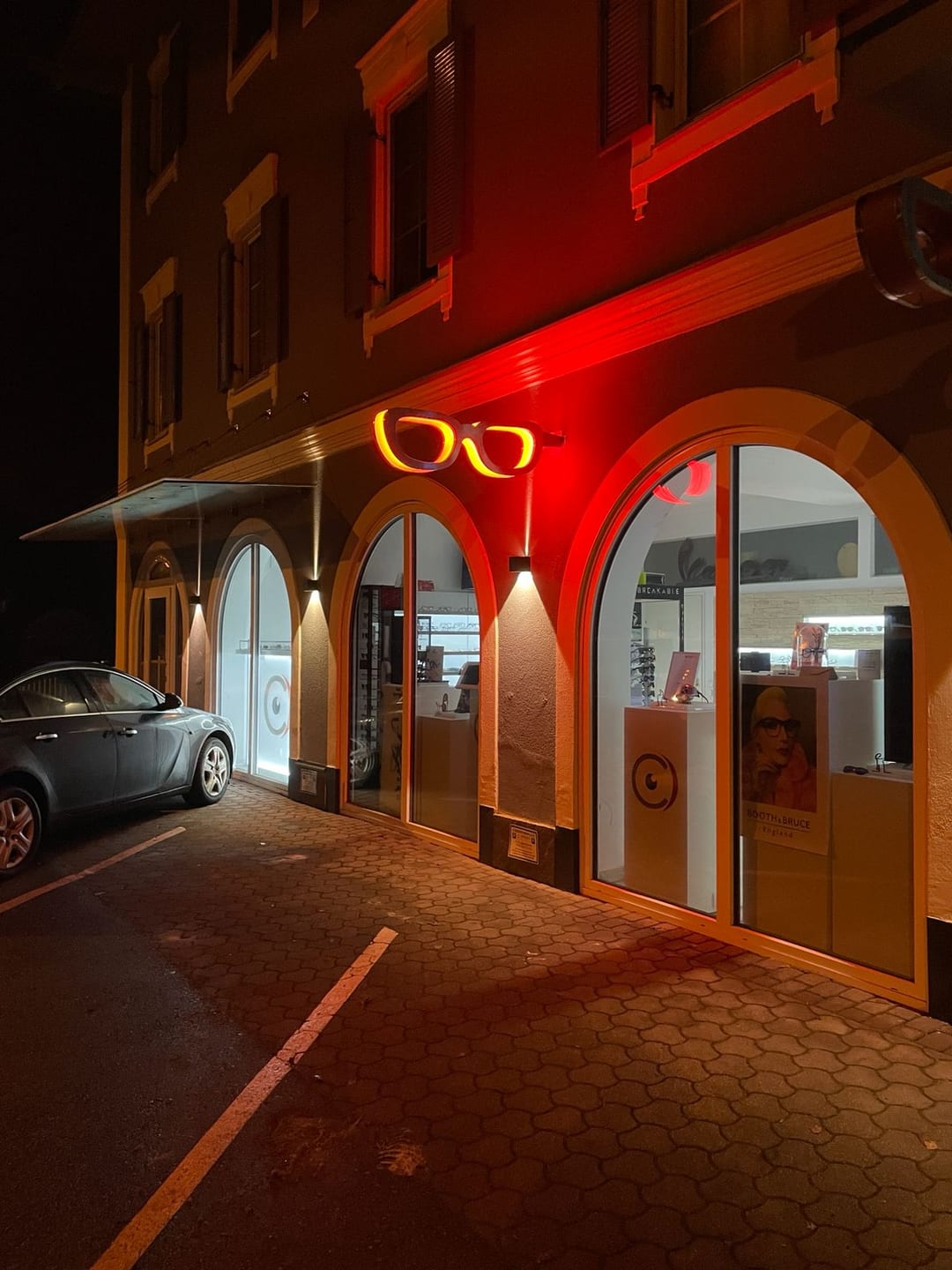 Leuchtende Brille als Ausleger an einer roten Fassade bei Nacht.