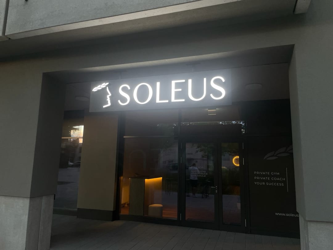 Weiß leuchtender Schriftzug „SOLEUS“ mit Logo auf einem Trägerkasten über dem Eingang bei Dämmerung.